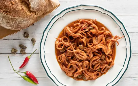Un piatto invitante di totani in umido, con capperi e peperoncino, accanto a un pane rustico.