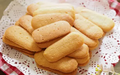 Un mucchio di biscotti a forma di lingue di gatto, dorati e friabili, disposti su una tovaglietta di pizzo bianco.