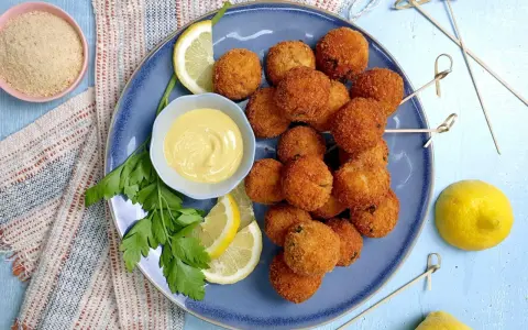 Piatto di polpette di tonno dorate e croccanti, servite con spicchi di limone, prezzemolo e una ciotolina di salsa gialla.