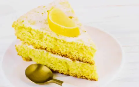 Fetta di soffice caprese al limone, spolverata di zucchero a velo e guarnita con una fetta di limone. Un cucchiaino dorato attende.