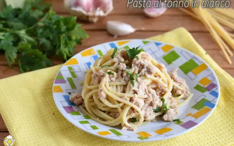 Un piatto di pasta al tonno in bianco, condita con tonno e prezzemolo, su un piatto colorato. Sullo sfondo, aglio e prezzemolo fresco.