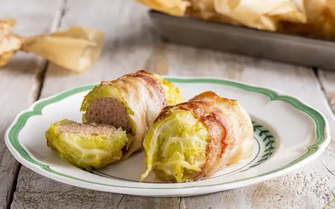 Deliziosi involtini di verza con ripieno di carne, avvolti in pancetta croccante, serviti su un piatto bianco con bordo verde.