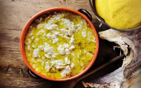 Piatto fumante di baccalà alla vicentina, con polenta gialla e stoccafisso essiccato sullo sfondo.