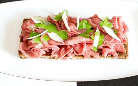 Delicato carpaccio di manzo su crostino integrale, guarnito con crema verde, scaglie di parmigiano e germogli freschi.