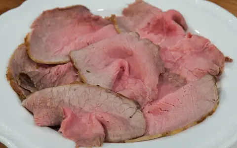 Fette sottili di arrosto di manzo rosa e succulento, servite su un piatto bianco.