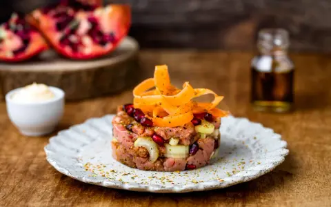 Tartare di manzo con melograno e carote, servita su un piatto rustico. Sullo sfondo, spicchi di melograno e una bottiglietta di condimento.