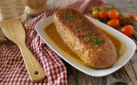 Polpettone in padella dorato e succulento, guarnito con erba cipollina, pronto per essere servito. Accanto, una spatola in legno e pomodorini freschi.