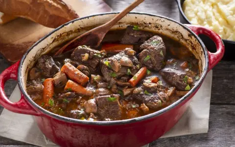 Un invitante spezzatino di manzo in un tegame rosso, con carote, funghi e un mestolo di legno. Accanto, pane rustico e purè di patate.