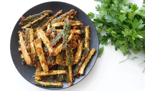 Zucchine gratinate in padella, dorate e croccanti, servite su un piatto nero con un ciuffo di prezzemolo fresco.