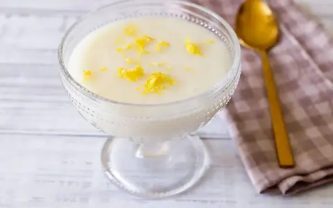 Deliziosa crema di riso in una coppa di vetro, guarnita con scorza di limone. Accanto, un cucchiaio dorato e un tovagliolo a quadretti.