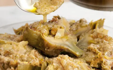 Un cucchiaio versa un sugo dorato su un piatto di carciofi alla romana, conditi con erbe aromatiche e pane croccante.