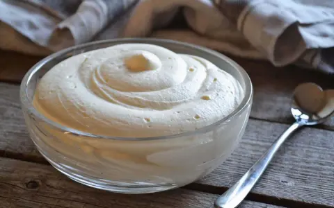 Una ciotola di vetro piena di soffice crema al mascarpone, con una spirale perfetta e una cucchiaio d'argento accanto.