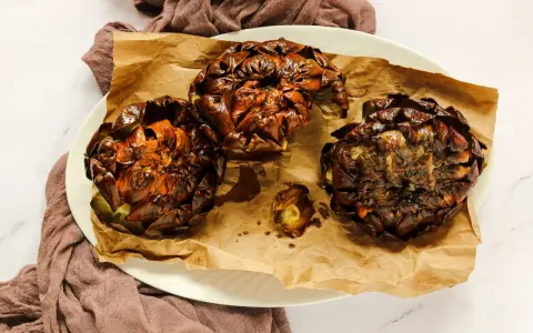 Tre carciofi alla giudia dorati e croccanti, serviti su carta da forno marrone, su un piatto bianco.