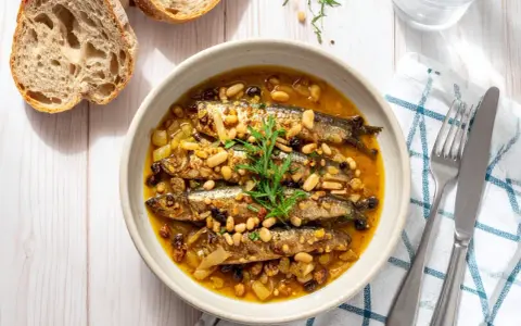 Sarde in saor in una ciotola, con pinoli, uvetta e cipolle. Accanto, pane rustico e posate.