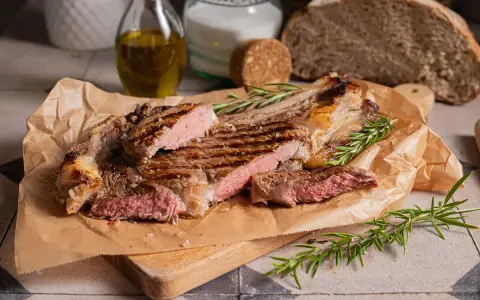Succulenta costata di manzo alla griglia, tagliata a fette, con rosmarino fresco e sale grosso. Sullo sfondo, pane rustico e olio d'oliva.
