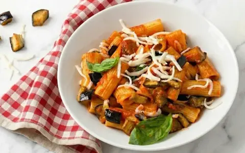 Un piatto di pasta alla norma fumante, con melanzane fritte, pomodoro e ricotta salata grattugiata, guarnito con basilico fresco.