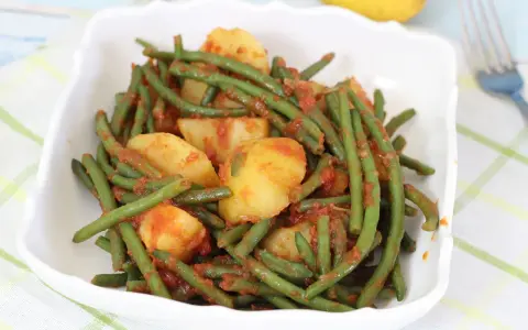 Un piatto di fagiolini in umido con patate, conditi con un sugo rosso saporito, servito in una ciotola bianca.