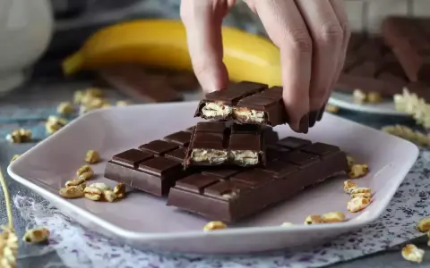 A hand breaks a chocolate bar filled with puffed rice and nuts on a pink plate.