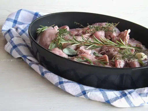 Pezzi di coniglio crudo in una teglia, pronti per essere cucinati al forno con rosmarino, salvia e aglio.