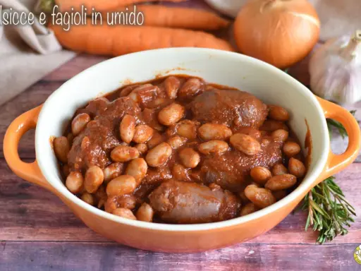 Un saporito piatto di salsiccia e fagioli in umido, servito in una ciotola arancione, con carote, cipolla e aglio sullo sfondo.