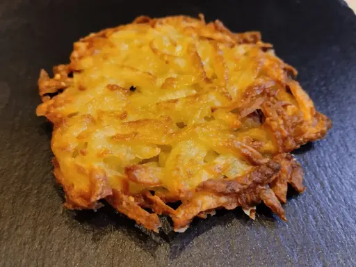 Un croccante rosti di patate dorato, con bordi leggermente bruciacchiati, adagiato su una superficie scura.
