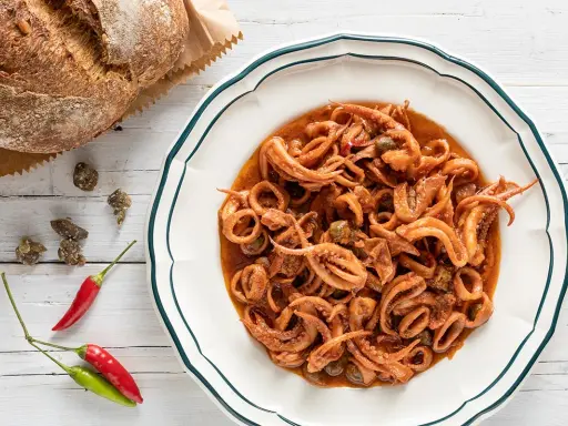 Un piatto invitante di totani in umido, con capperi e peperoncino, accanto a un pane rustico.