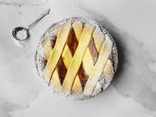 Pastiera napoletana pronta, cosparsa di zucchero a velo. Un cucchiaino d'argento è pronto per decorare, mostrando come si fa la pastiera.