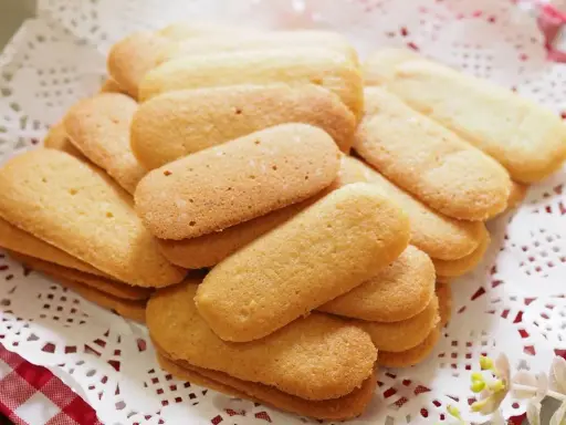 Un mucchio di biscotti a forma di lingue di gatto, dorati e friabili, disposti su una tovaglietta di pizzo bianco.