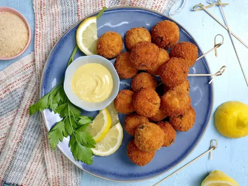 Piatto di polpette di tonno dorate e croccanti, servite con spicchi di limone, prezzemolo e una ciotolina di salsa gialla.