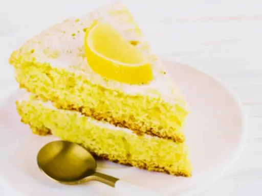 Fetta di soffice caprese al limone, spolverata di zucchero a velo e guarnita con una fetta di limone. Un cucchiaino dorato attende.