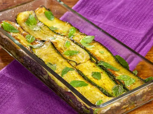 Zucchine alla scapece dorate e profumate alla menta, servite in una pirofila di vetro su un tovagliolo viola.