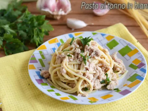 Un piatto di pasta al tonno in bianco, condita con tonno e prezzemolo, su un piatto colorato. Sullo sfondo, aglio e prezzemolo fresco.