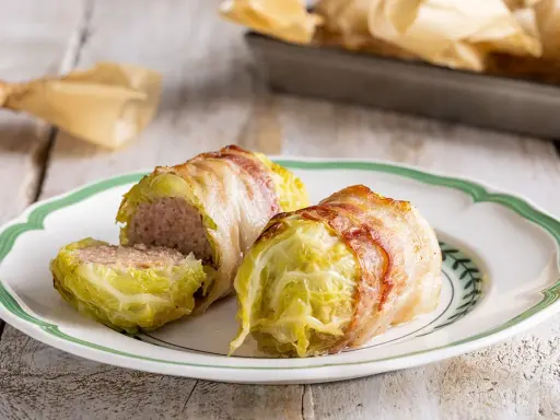Deliziosi involtini di verza con ripieno di carne, avvolti in pancetta croccante, serviti su un piatto bianco con bordo verde.