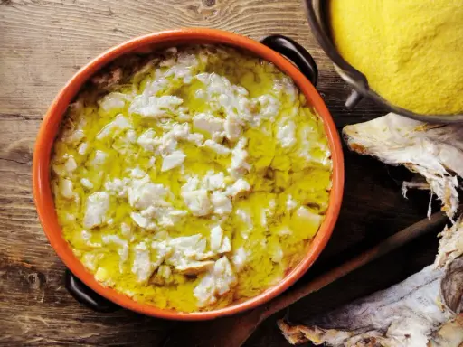 Piatto fumante di baccalà alla vicentina, con polenta gialla e stoccafisso essiccato sullo sfondo.