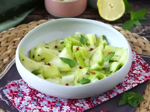 Delicate ribbons of zucchine marinate crude, tossed with fresh mint and pink peppercorns, served in a white bowl.