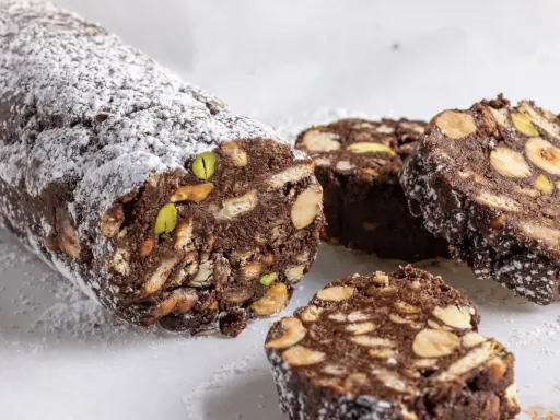 Salame di cioccolato senza uova, ricco di biscotti, nocciole e pistacchi, spolverato di zucchero a velo.