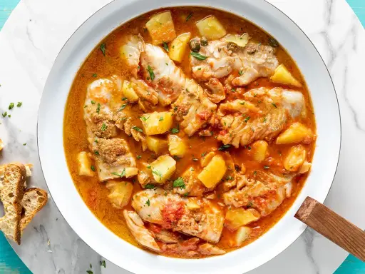 Un invitante merluzzo in umido con patate, pomodoro e capperi, guarnito con prezzemolo fresco e accompagnato da fette di pane croccante.