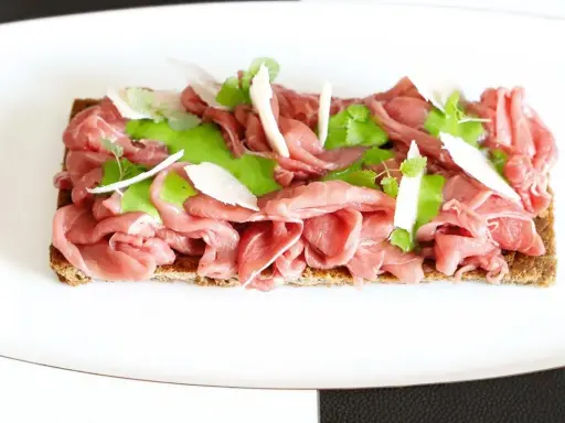 Delicato carpaccio di manzo su crostino integrale, guarnito con crema verde, scaglie di parmigiano e germogli freschi.