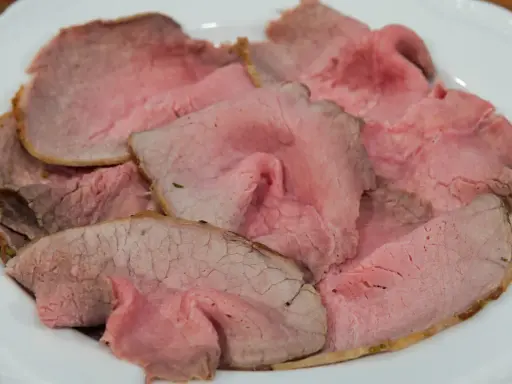 Fette sottili di arrosto di manzo rosa e succulento, servite su un piatto bianco.