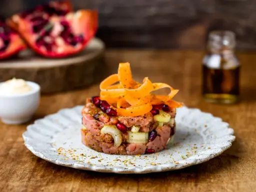 Tartare di manzo con melograno e carote, servita su un piatto rustico. Sullo sfondo, spicchi di melograno e una bottiglietta di condimento.