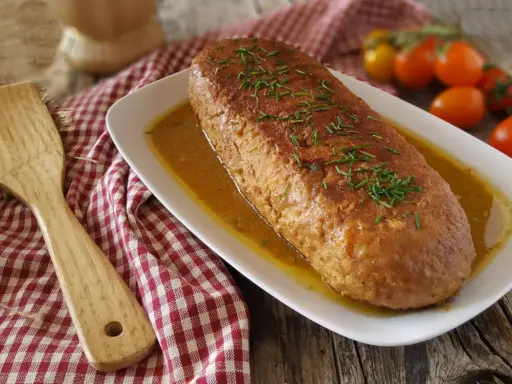 Polpettone in padella dorato e succulento, guarnito con erba cipollina, pronto per essere servito. Accanto, una spatola in legno e pomodorini freschi.