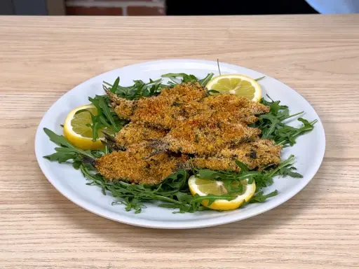 Piatto di alici al forno dorate e croccanti, servite su un letto di rucola fresca con spicchi di limone.