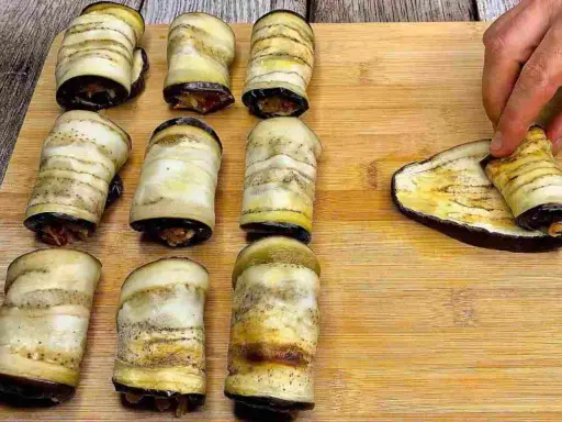 Involtini di melanzane pronti per essere infornati. Una mano sta arrotolando l'ultima fetta di melanzana grigliata.