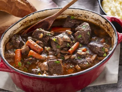 Un invitante spezzatino di manzo in un tegame rosso, con carote, funghi e un mestolo di legno. Accanto, pane rustico e purè di patate.