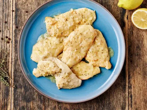Piatto di pollo al limone su un piatto blu, con limone e rosmarino sullo sfondo.