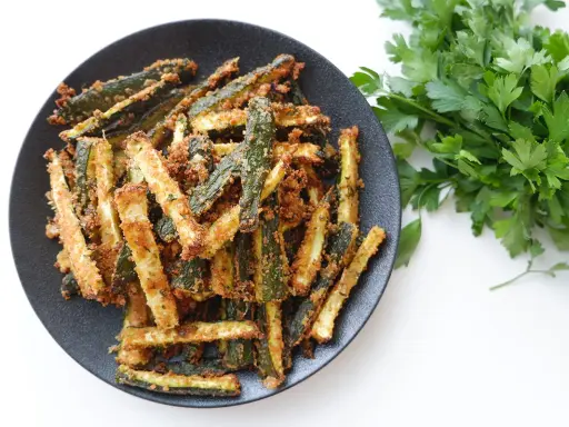 Zucchine gratinate in padella, dorate e croccanti, servite su un piatto nero con un ciuffo di prezzemolo fresco.