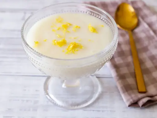 Deliziosa crema di riso in una coppa di vetro, guarnita con scorza di limone. Accanto, un cucchiaio dorato e un tovagliolo a quadretti.