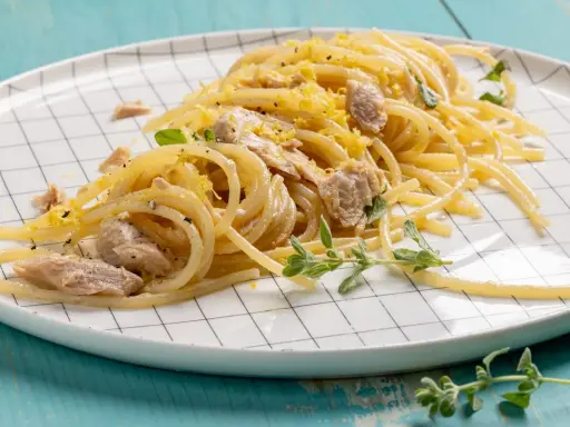 Un piatto di pasta tonno e limone, condita con scorza di limone grattugiata e erbe aromatiche fresche, servita su un piatto a scacchi bianchi.