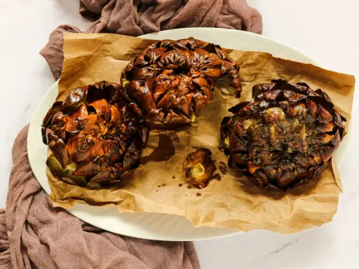Tre carciofi alla giudia dorati e croccanti, serviti su carta da forno marrone, su un piatto bianco.
