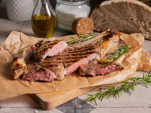 Succulenta costata di manzo alla griglia, tagliata a fette, con rosmarino fresco e sale grosso. Sullo sfondo, pane rustico e olio d'oliva.